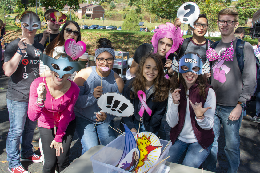 A group of students pose with photo booth props, including superhero garb and pink breast cancer awareness cutouts