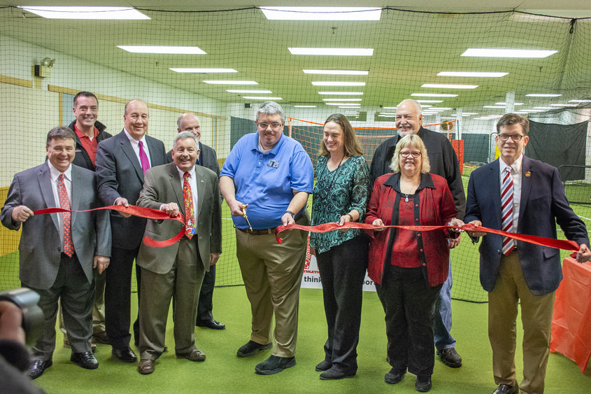 Several people stand on turf grass in a line while holding a red ribbon. Joe Medica stands in the center holding scissors to cut the ribbon.