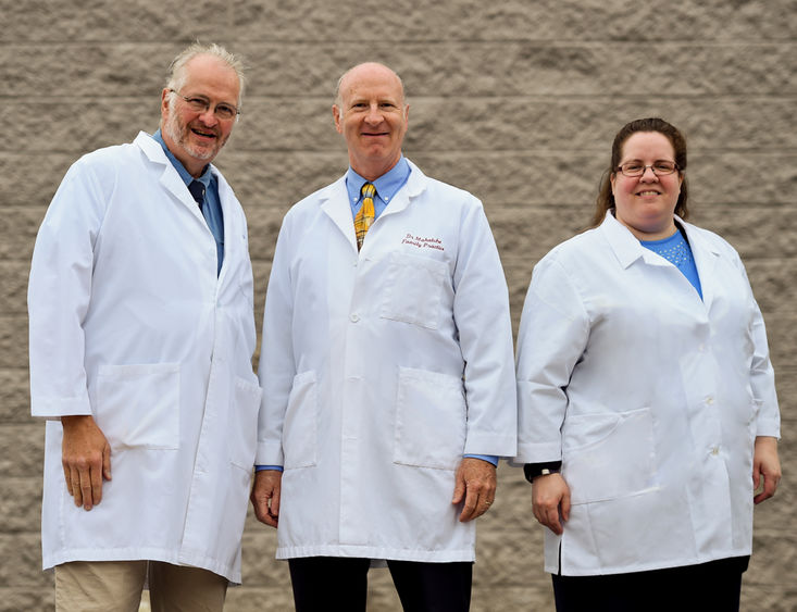 Three people -- two men and one woman -- stand in front of a beige, textured wall, posing for a photo. They're each wearing white clinical jackets.
