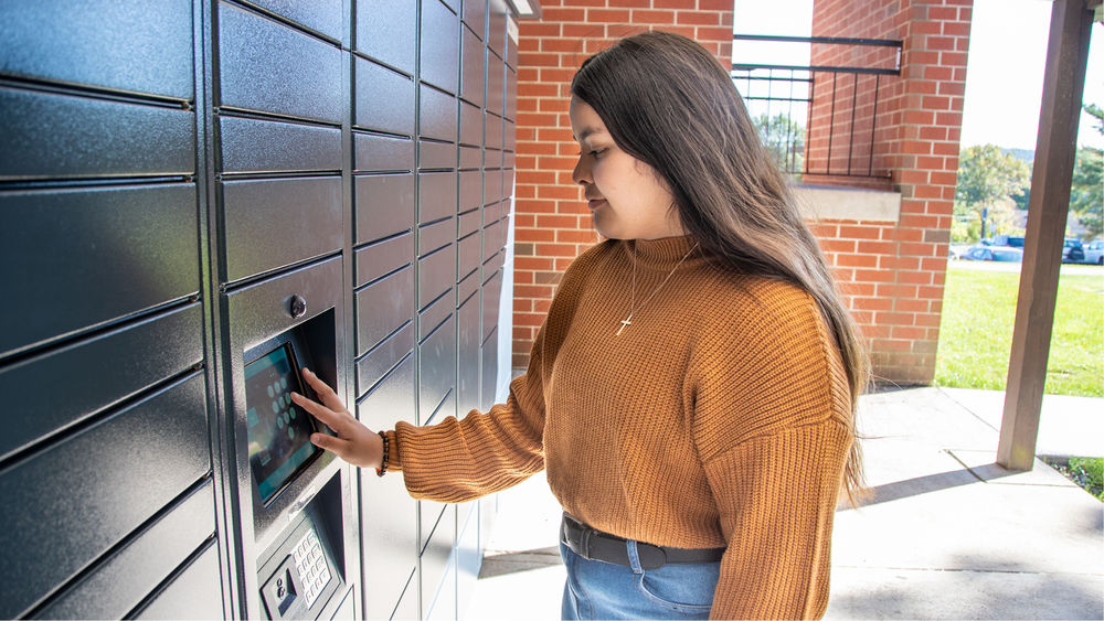 Student with long brown hair wears a burnt orange sweater and jeans as she types on a digital keyboard at navy lockers