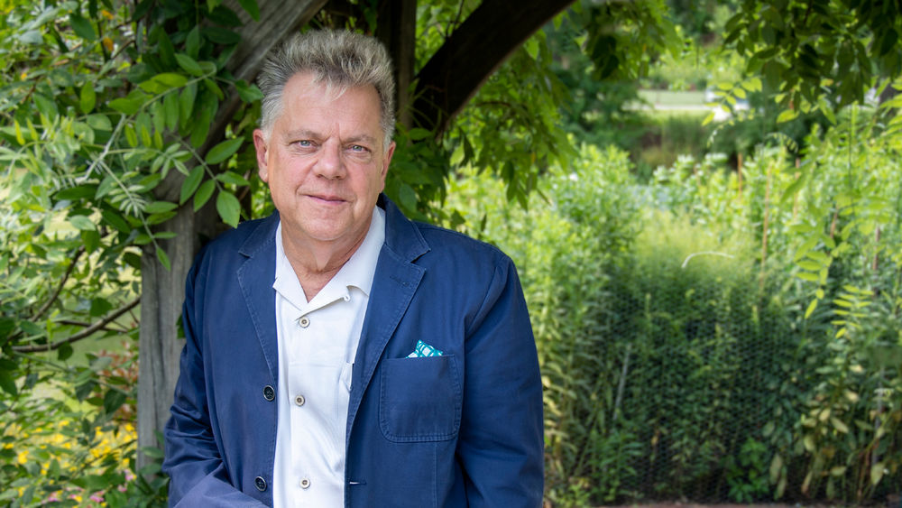 Man wearing white button-down shirt and blue jacket leans against a wooden bower in garden