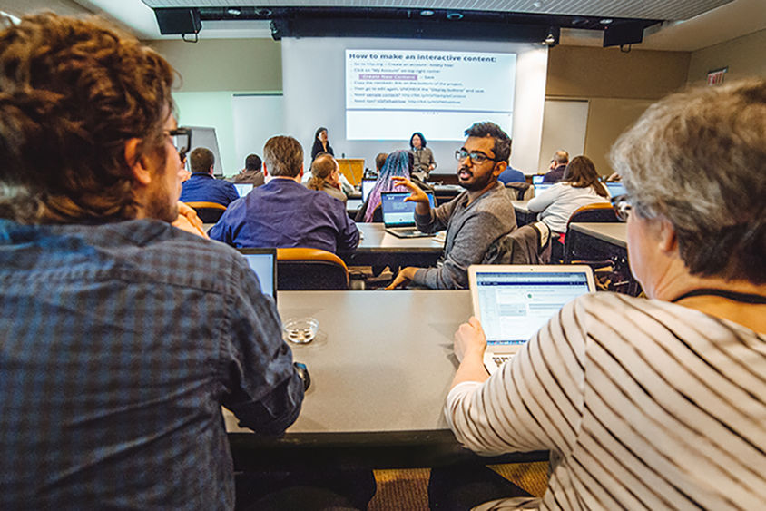 A group of people seated with laptops looking  at two presenters standing in the front of the room with an overhead presentation on the screen