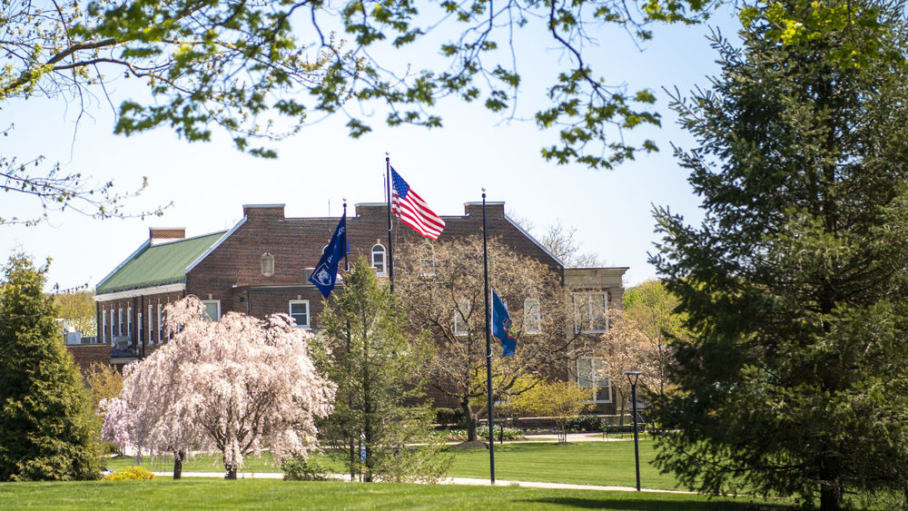 U.S., Pennsylvania, and Penn State Schuylkill flags fly in the wind over a large grassy field with a brick building in the background.