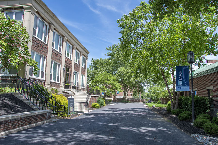 Two story brick building on left hand of photo with concrete pathway lined by trees