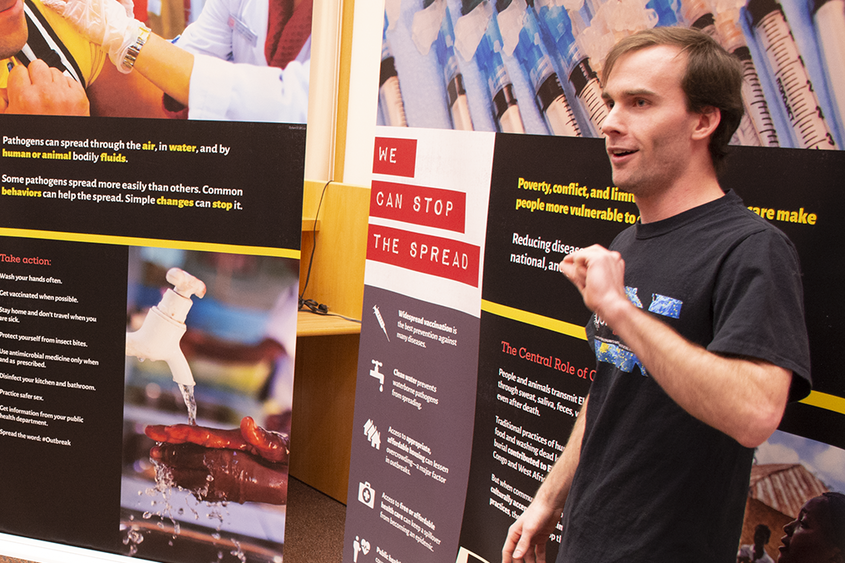 A man wearing a black t-shirt stands in front of signage about viral outbreaks