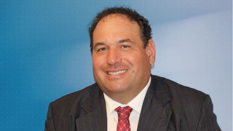 Headshot of Jonathan Kukta, Penn State Schuylkill's director of business services, wearing a gray suit, white shirt, and red tie, and standing in front of a blue background