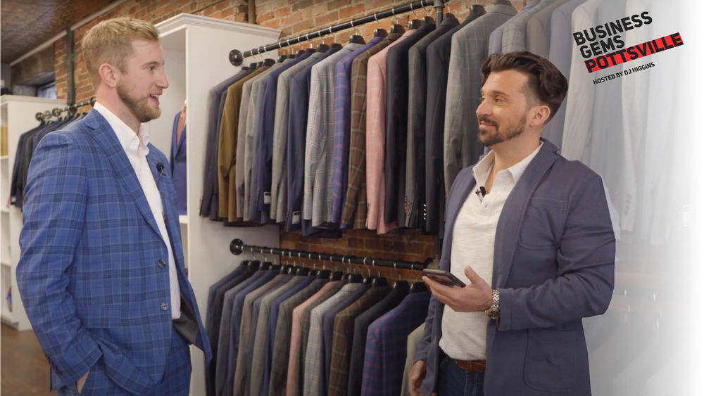 Two men wearing suits talk in a men's clothing boutique as they stand in front of sports jackets with the "Business Gems Pottsville" logo on the right