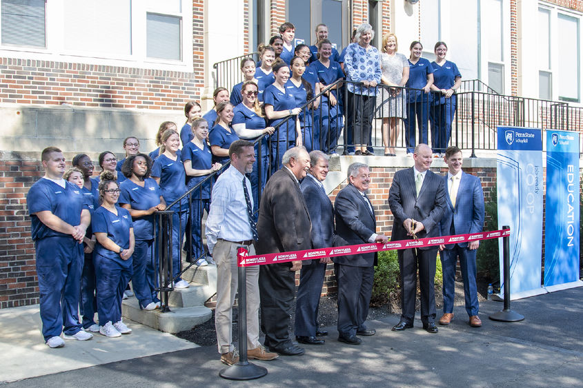 Students wearing dark blue scrubs line stairs in front of a brick building. Six men wearing suits stand in front of them while holding a red ribbon to be cut by large gold scissors.