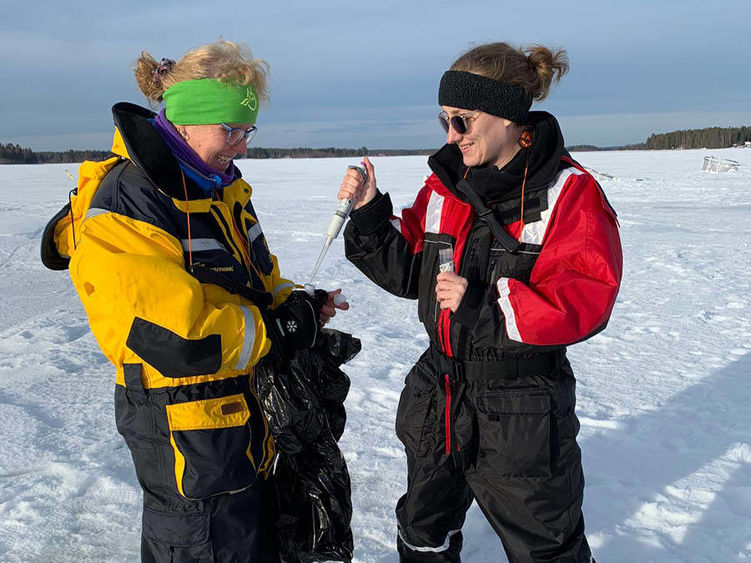 Two women wear winter weather gear and sunglasses while using a micropipette while walking on a frozen lake