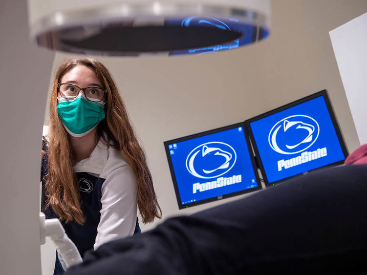Girl wearing surgical mask operates radiological sciences equipment with two computer monitors in the background with Penn State athletics logo