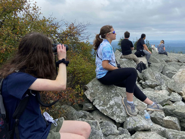 Students wearing active wear sit atop a mountain peering through binoculars.