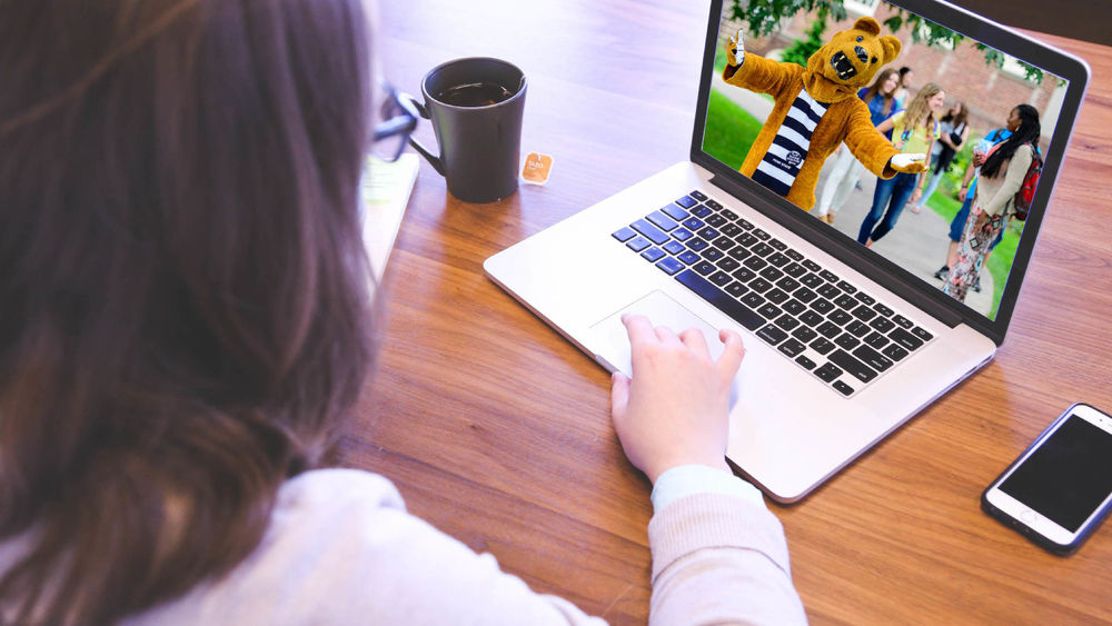 A student sits at a desk with a mug of hot tea, a cell phone, and a computer with Penn State's Nittany Lion mascot welcoming her on the screen.